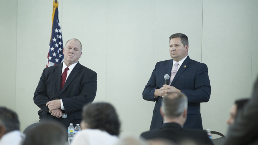 Two men in suits standing in front of room, white wall, with American flag at side