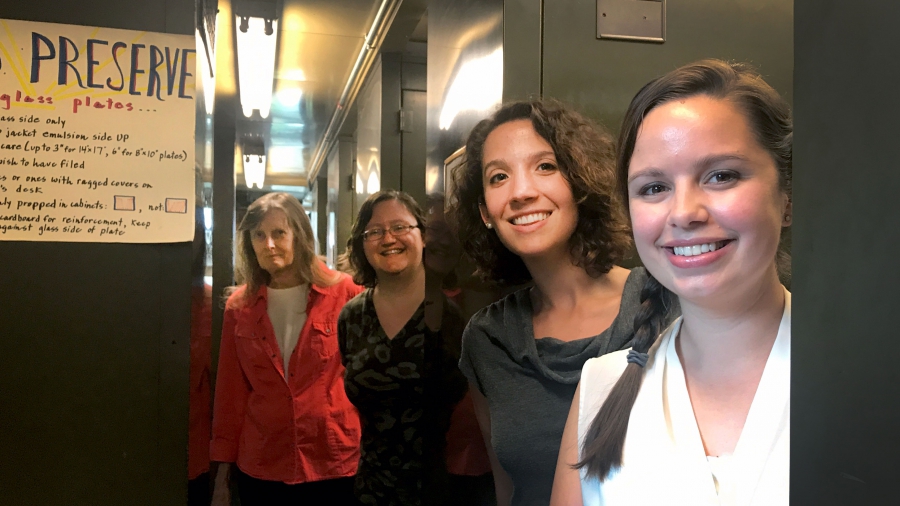 A group of four women pose in the Harvard plate stacks, surrounded by tall metal cabinets. A paper sign to their left says "preserve the glass."