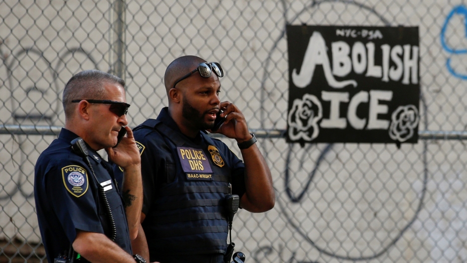 Department of Homeland Security officers watch members of Occupy ICE, a group set on disrupting Immigration and Customs Enforcement (ICE) operations, outside the ICE offices in New York City, June 25, 2018. Two uniformed Department of Homeland Security officers are shown looking off to the right of the frame while talking on their walkie-talkies.