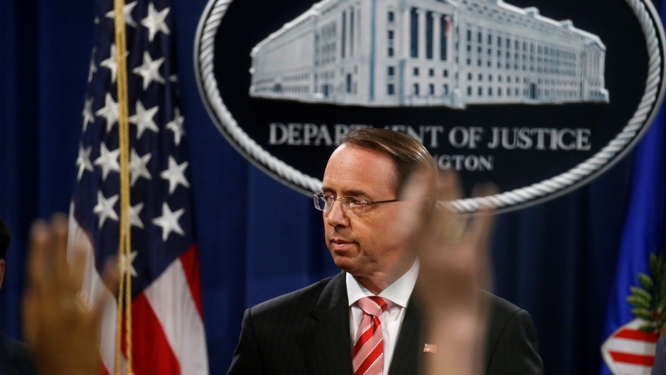 Deputy US Attorney General Rod Rosenstein takes questions after announcing grand jury indictments of 12 Russian intelligence officers in special counsel Robert Mueller's Russia investigation, during a news conference at the Justice Department in Washingto Deputy US Attorney General Rod Rosenstein is seen in front of an emblem of the US Justice Department with reporter's hands raised in the near ground.