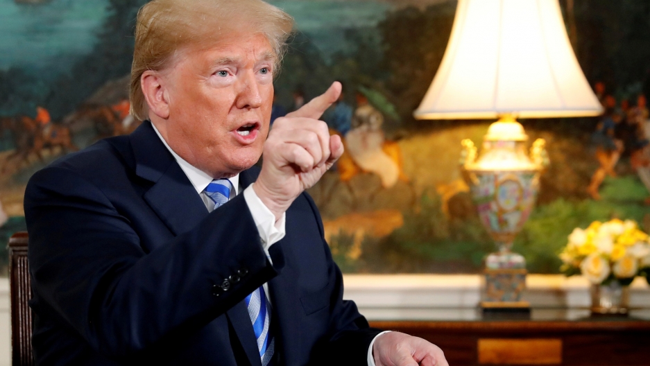 US President Donald Trump speaks to reporters after signing a proclamation declaring his intention to withdraw from the JCPOA Iran nuclear agreement. US President Donald Trump is shown sitting at this desk with his right hand pointing.