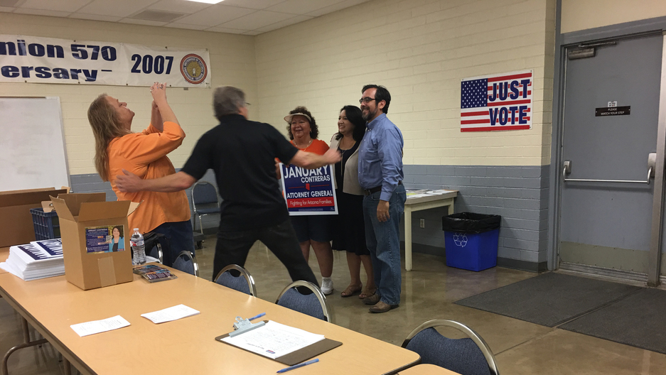 People stand in auditorium-like room in front of folding table with a clipboard on it, surrounding a woman, in support