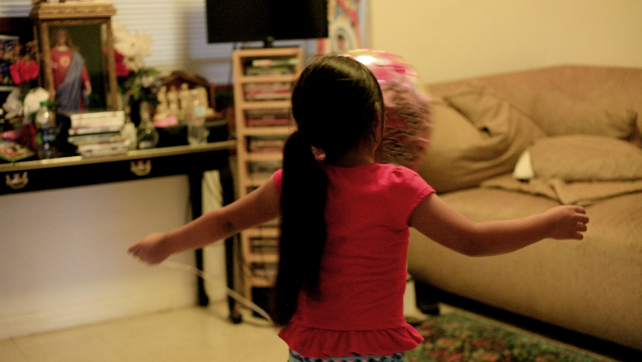 Young girl, shown from behind, with arms outstreched, in room in home