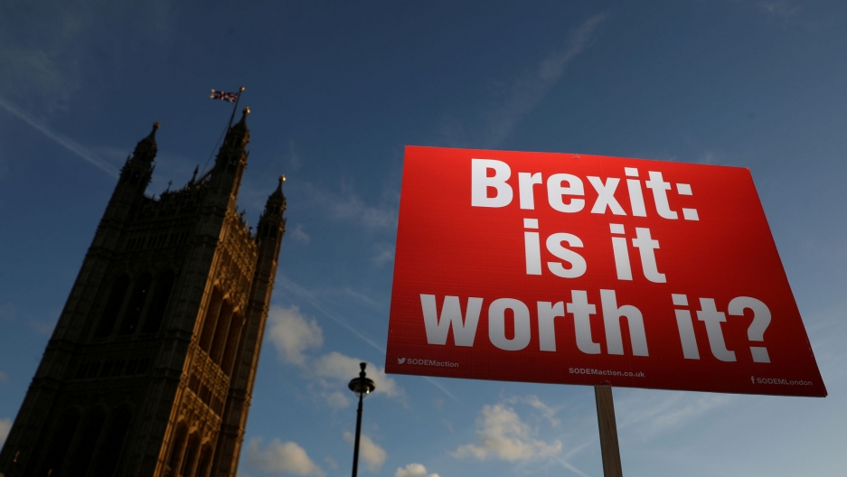 An anti-Brexit demonstrator holds a placard opposite the Houses of Parliament in London. A red placard with white lettering reading, "Brexit: is it worth it?" is shown in the foreground with the Houses of Parliament in the background.