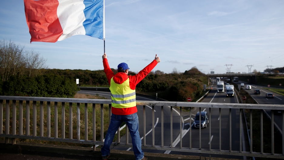 A man wearing a yellow vest and holding a French flag stands on a bridge near the Nantes Atlantique Airport. A man wearing a yellow vest and holding a French flag stands on a bridge with his arms in the air.
