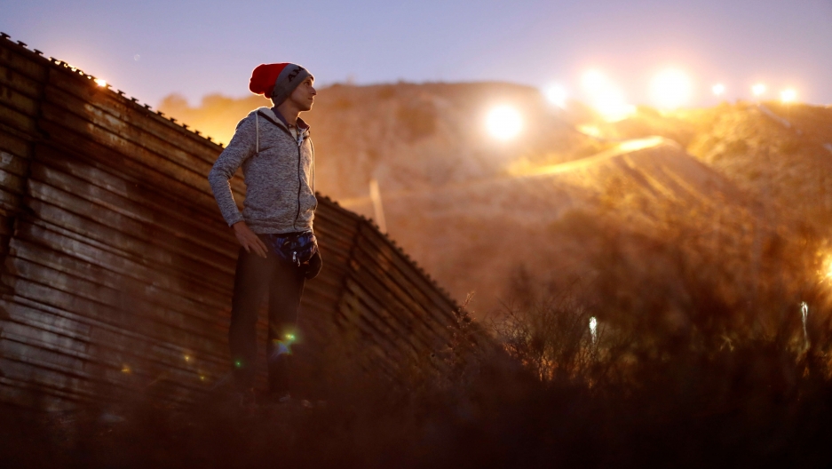 A migrant from Honduras stands in front of the border wall with the sun shining behind him 