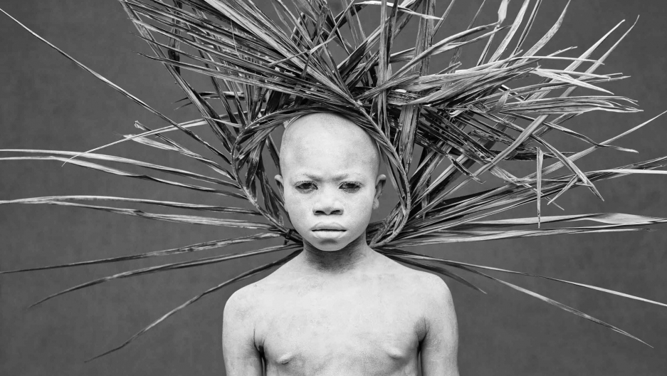 A boy whose body is painted white wears a crown of wooden sticks that looks like a sunburst behind his head poses for a photo