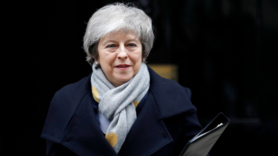 Britain's Prime Minister Theresa May is shown wearing a grey scarf and blue overcoat with a binder in her left hand.