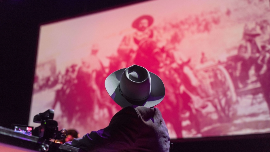 a performer wearing a hat looks at the giant projection of Pancho Villa on the stage film screen