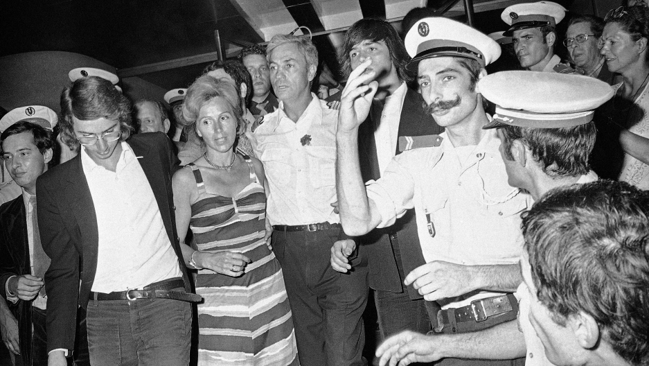 A black and white photo showing a man in a pilot's uniform with a mustache looking at a camera among a crowd of people.  