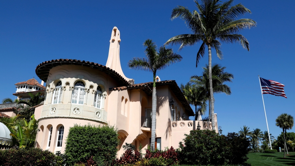 US President Donald Trump's Mar-a-Lago estate in Palm Beach, Florida. The Mar-a-Lago estate is shown in a medium shot photograph with peach-colored walls and a US flag flying to the right.