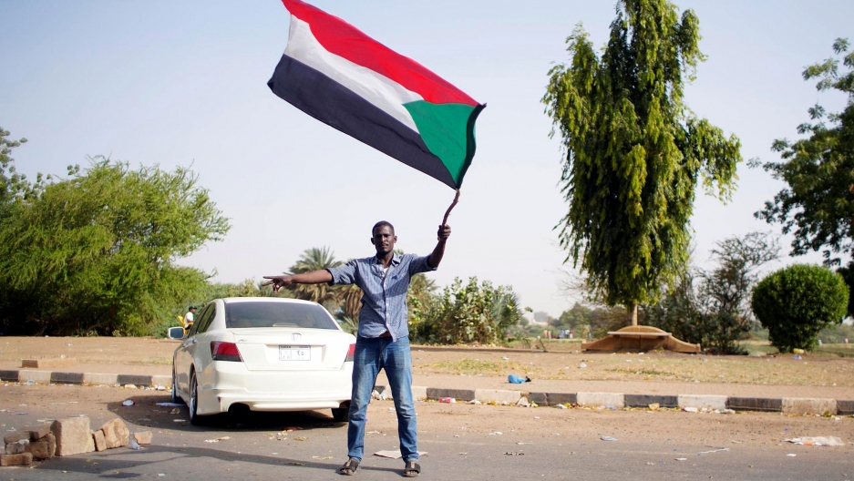 A Sudanese demonstrator waves a national flag as he arrives to protest in Khartoum. A man is shown wearing sandals, jeans and a button down shirt while waving the Sudanese flag.