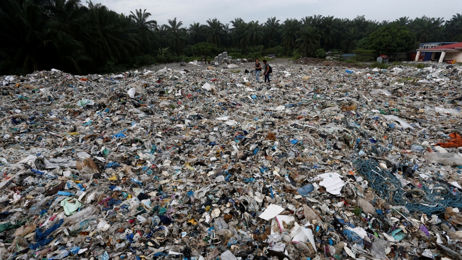Plastic waste are piled outside an illegal recycling factory in Jenjarom, Kuala Langat, Malaysia, 2018. Plastic waste is shown covering almost the entire ground area of the photo with a slim green tree line along the top of the frame.