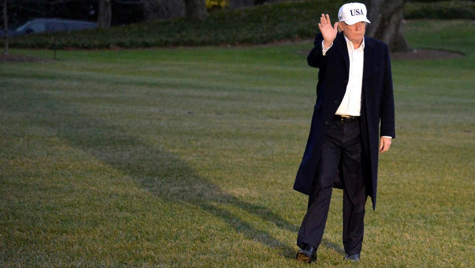 President Trump, wearing a white baseball hat with the letters U.S.A. embroidered, waves to cameras while walking on the White House lawn