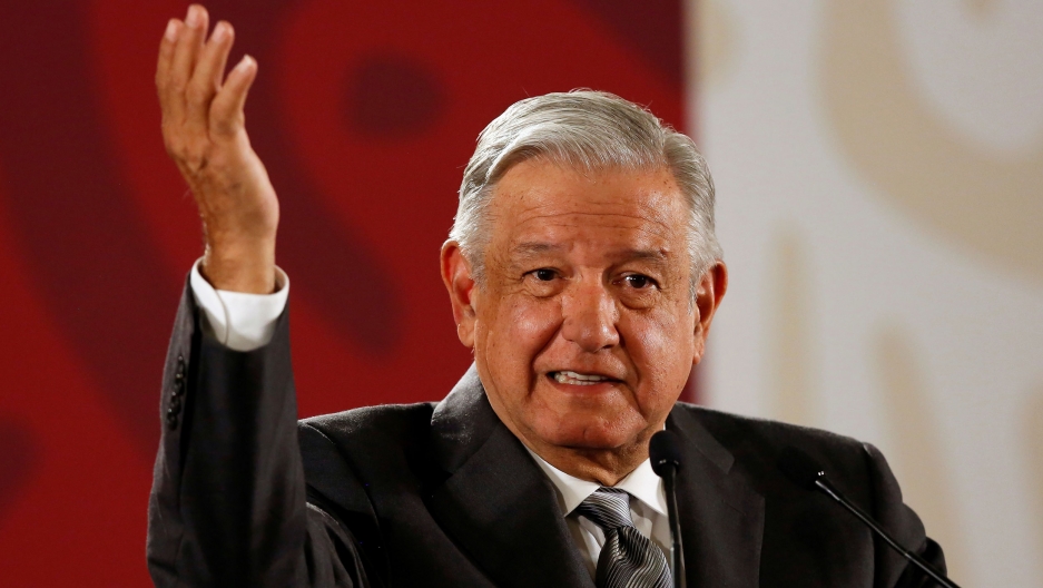 Mexico's President Andrés Manuel López Obrador gestures. Mexico's President Andrés Manuel López Obrador is shown with his right hand raised in the air and a red and white background.