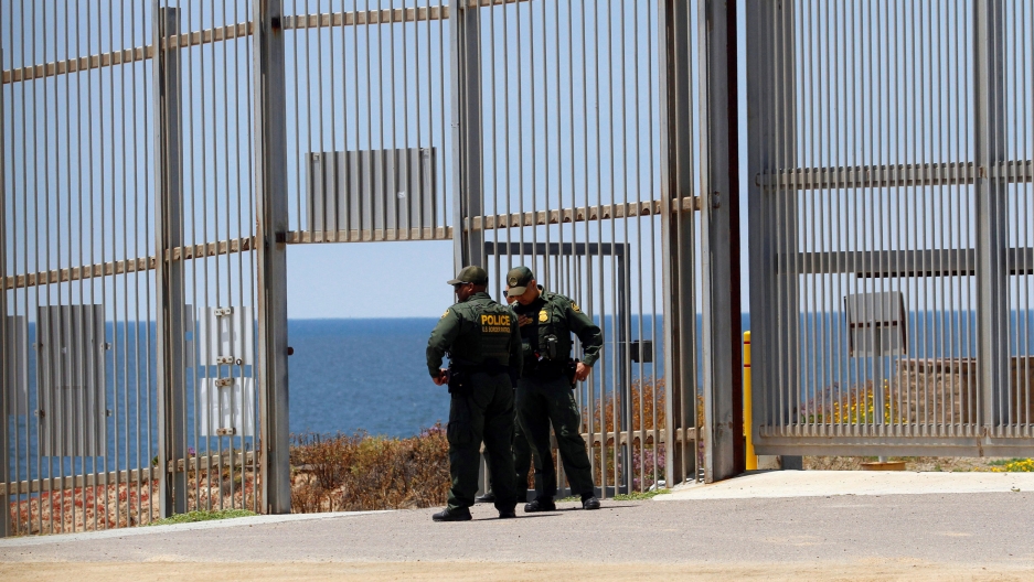 US Border Patrol agent are seen in the border fence between the United States and Mexico as photographed from Tijuana, Mexico, June 1, 2019. Two US Border Patrol agent are seen standing in uniform with their hands on their hips.