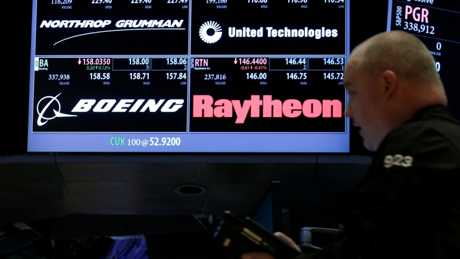 Trader passes by a screen that displaying Northrop Grumman and United Technologies who announced merger plans on Sunday. A man is shown in soft focus in the nearground with a stock trading electronic board in the background.