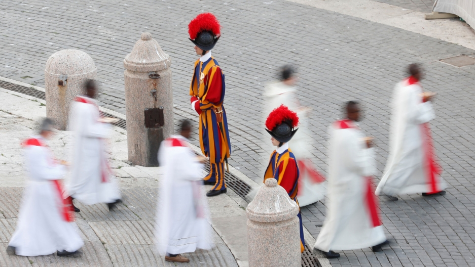 Priests walk past Swiss guards during a vigil led by Pope Francis at the Vatican. Several Catholic priests wearing white robes are shown walking past two guards in traditional striped uniforms.