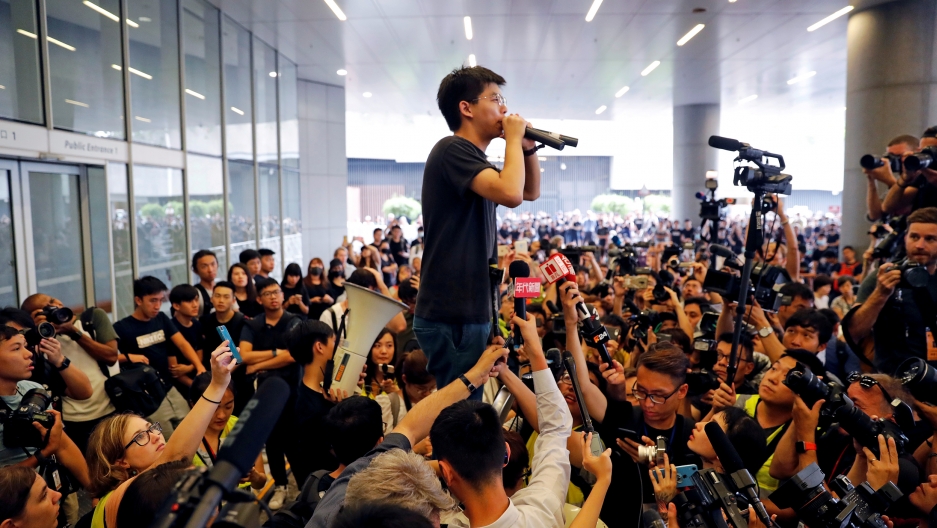 A man holds a microphone and is surrounded by dozens and dozens of news media and others taking photos and video