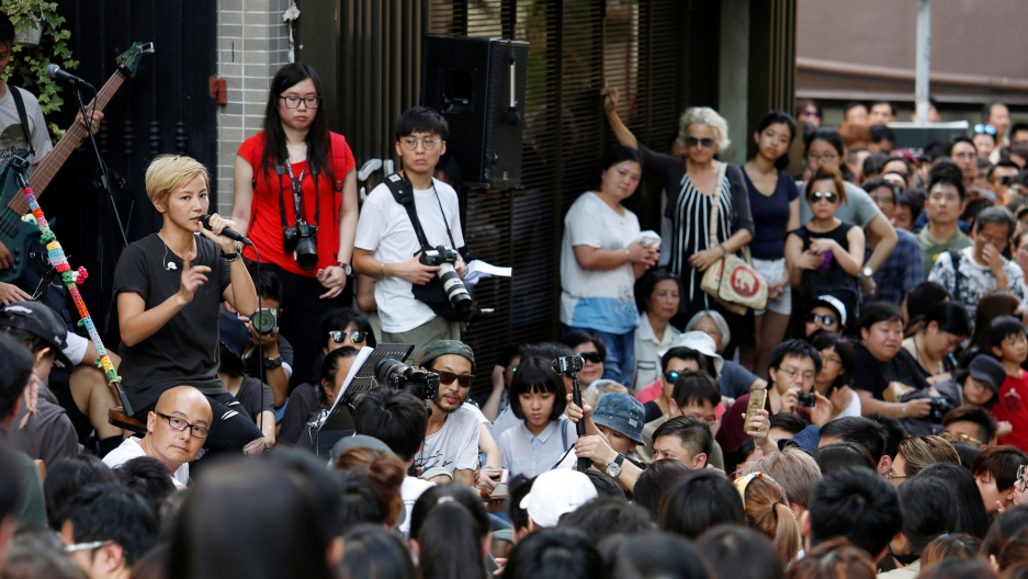 Thousands of people cram the streets of Hong Kong, walking off a freeway exit. The buildings of the city are behind them