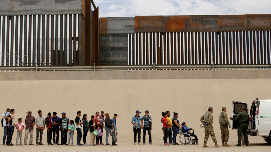 Members of the Border Patrol and US Military help a migrant woman in a wheelchair as she and others migrants crossed the border between Mexico and the US to request political asylum, as seen from Ciudad Juarez, Mexico, July 6, 2019. A line of migrants that includes young and older people along with a person in a wheelchair are shown standing next to the large border fence.