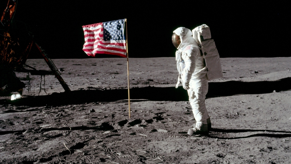 Astronaut Buzz Aldrin poses for a photograph beside the United States flag on the moon. Buzz Aldrin stands in his full astronaut suit facing the United States flag.