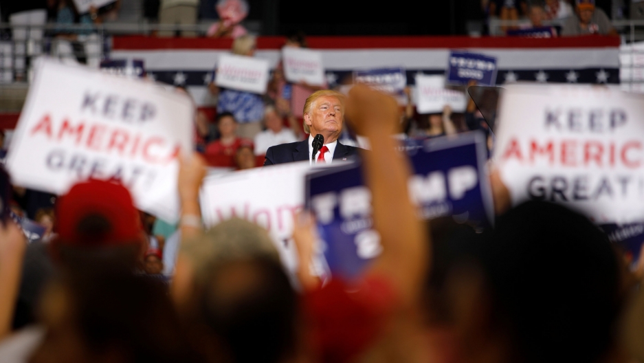 US President Donald Trump pauses while speaking at a campaign rally in Greenville, North Carolina, July 17, 2019. A large crowd of people are shown in soft focus holding placards with Donald Trump in center frame in focus.