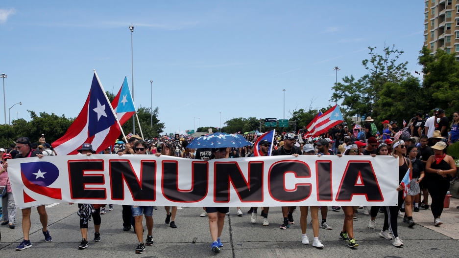 People hold a banner that reads "Resign" during a protest calling for the resignation of Governor Ricardo Rosselló in San Juan, Puerto Rico, July 22, 2019. A large group of people are shown walking down the street and holding a long banner with the word "Renuncia" printed on it.