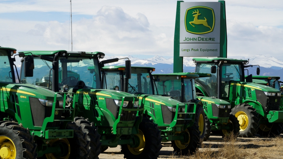 John Deere tractors are seen for sale in Colorado. Several green John Deere trackers are shown in a row with a sign for the company in the background.