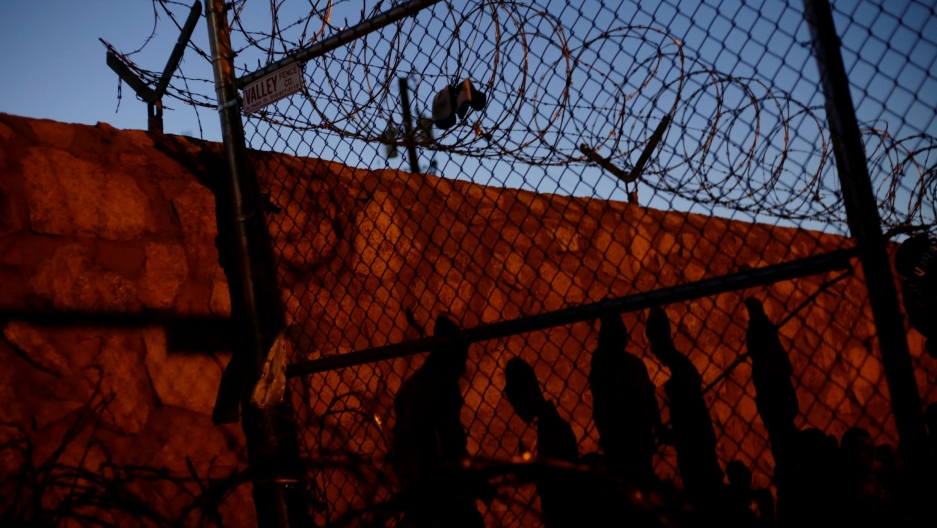 Central American migrants cast their shadows against a wall while inside an enclosure where they are being held by US Customs and Border Protection, after crossing the border between Mexico and the United States to request asylum, in El Paso, Texas. The shadows of five people are shown on a wall with a chainlink fence in the nearground.