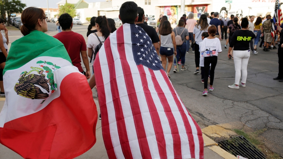 People with the Mexican flag and the US flag take part in a rally against hate a day after a mass shooting at a Walmart store, in El Paso, Texas, Aug. 4, 2019. Two people are shown walking away from the camera with a Mexican flag and a US flag draped over their shoulders like capes.
