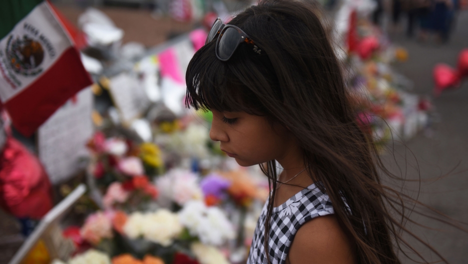 A young girl looks at a growing memorial three days after a mass shooting at a Walmart store in El Paso, Texas. Aug 6, 2019. A young girl is shown in a checkered dress with long dark hair and sunglasses, standing in front of a memorial.