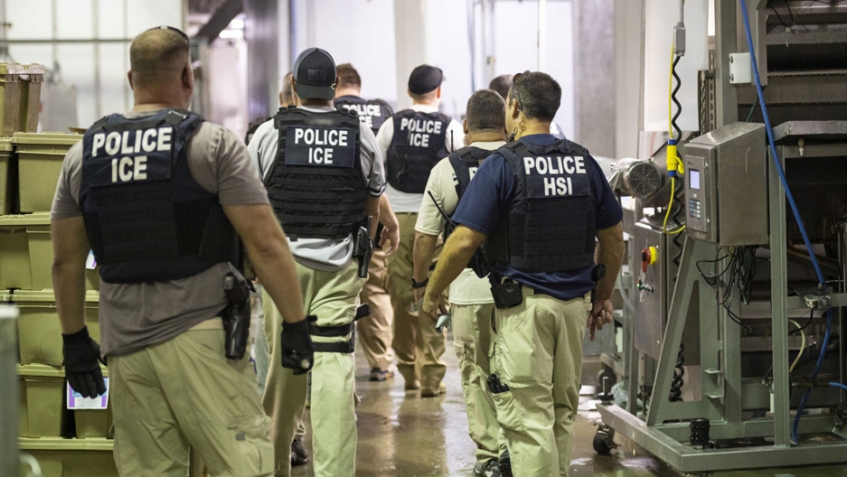 Officers from Immigration and Customs Enforcement look on after executing search warrants and making some arrests at an agricultural processing facility in Canton, Mississippi. Several ICE officers are shown walking away from the camera wearing bullet-proof vests and gun holsters on their sides.