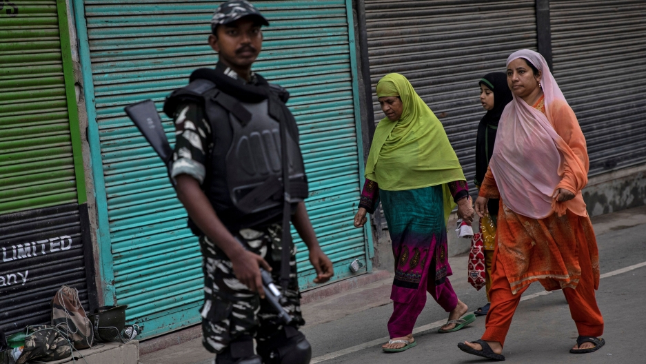 Kashmiri women walk past Indian security personnel during restrictions Aug. 11, 2019, after the special constitutional status for Kashmir was revoked on Aug. 5. Three women are shown in colorful attire and head coverings walking past an armed security official.