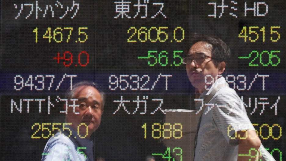 Passersby are reflected on a stock quotation board outside a brokerage in Tokyo, Japan. Two men are shown in a reflection of a stock market ticker wearing collared shirts