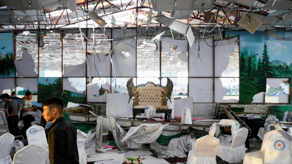 Workers of a wedding hall inspect after a blast in Kabul, Afghanistan August 18, 2019. ISIS claimed responsibility on Sunday for a suicide blast at a wedding reception that killed 63 people. A young man is show walking off to the left through the debris room filled with white-covered chaies and a ceiling falling down.
