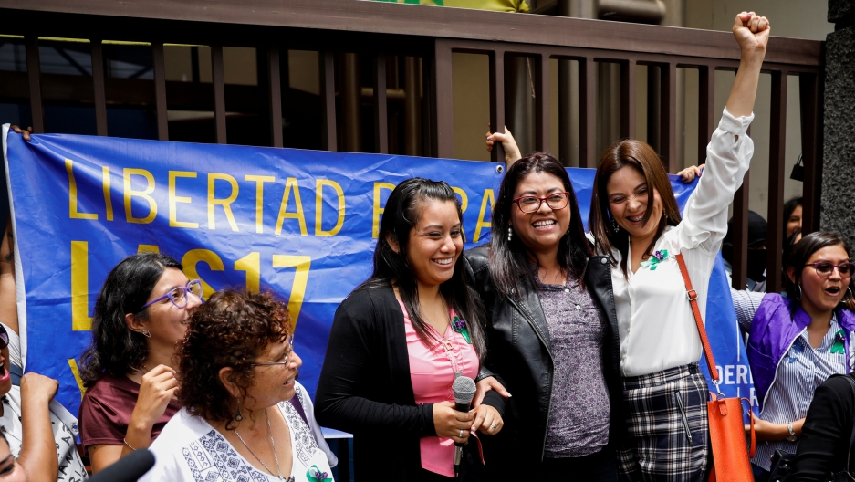Evelyn Hernández (Center), who was sentenced to 30 years in prison for a suspected abortion, talks to the media after being absolved at a hearing in Ciudad Delgado, El Salvador, Aug. 19, 2019. Evelyn Hernández is shown with a black sweater and pink shirt standing among several other women smiling.