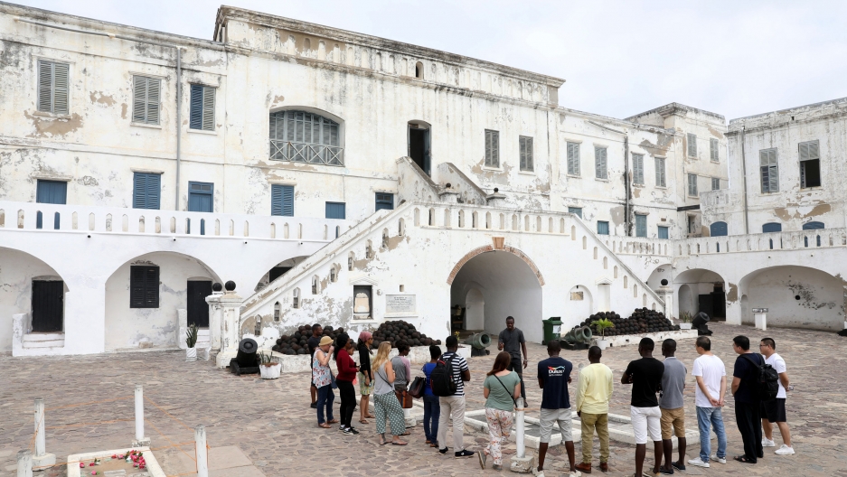 Tourists listen to a guide at the Cape Coast Slave Castle in Ghana, July 28, 2019. Cape Coast Castle was used as a slaving post from where slaves were bought and sold and then shipped to what would become the US and other countries. Several people are shown standing around a tour guide in front of the white facade of the Cape Coast Castle.