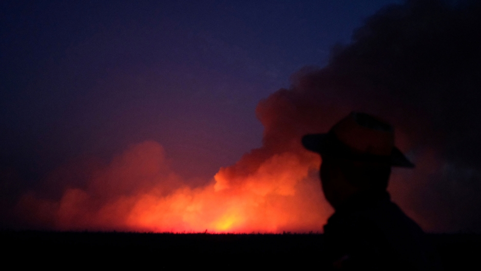 A man is seen standing in shadow with a burning tract of the Amazon jungle in Canarana, Brazil, seen in the distance, Aug. 26, 2019. A man wearing a have is shown in the near ground, black in shadow, with a brigh orange fire seen in the distance.