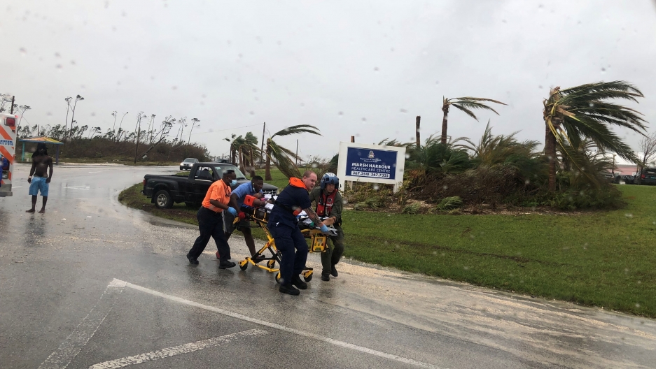 US Coast Guard personnel help medevac a patient during Hurricane Dorian in the Bahamas, Sept. 3, 2019. Several rescue workers are shown moving a yellow emergency stretcher down a road with winds blowing the trees in the background.