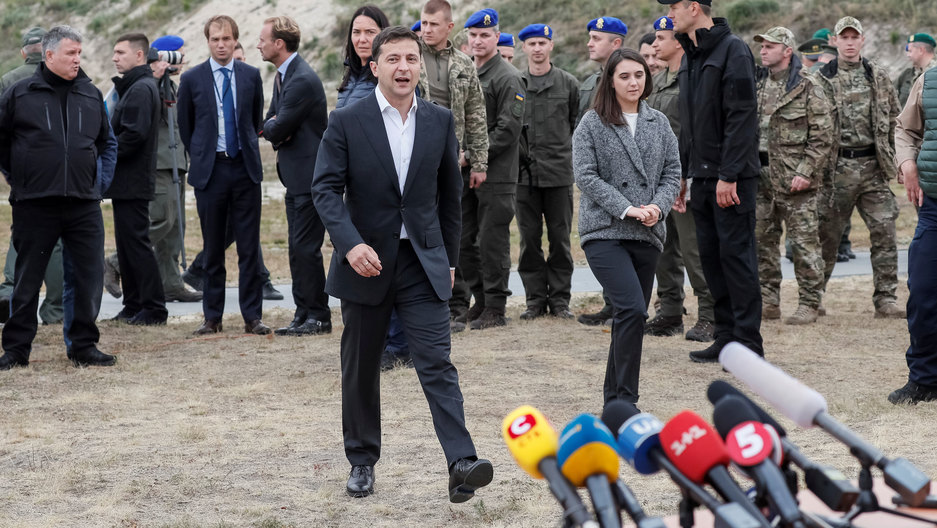 Ukrainian President Zelenskiy walks toward microphones before speaking with journalists near the village of Stare in Kyiv region, Ukraine, Sept. 30, 2019. Ukrainian President Zelenskiy is shown wearing a dark suite and walking toward several microphones in an outdoors setting.