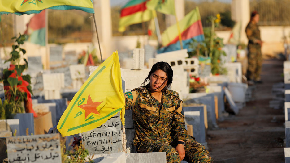 Fighters of Syrian Democratic Forces visit the graves of their late comrades at a cemetery in Kobani, Syria, 2017. A woman wearing military fatigues is shown sitting in a cemetary with several flags flying.