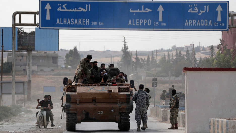 Turkey-backed Syrian rebel fighters sit on a military truck at the border town of Tel Abyad, Syria, Oct. 14, 2019. Several people are shown sitting on a military truck passing under a large blue road sign overhead.