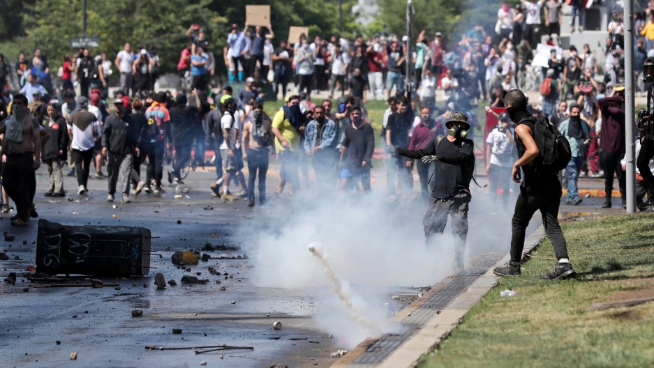 Demonstrators deflect a tear gas canister during a protest in Santiago, Chile, Oct 20, 2019. A crowd of demonstrators are shown amassed in the street with a tear gas canister bouncing in the middle of the frame.
