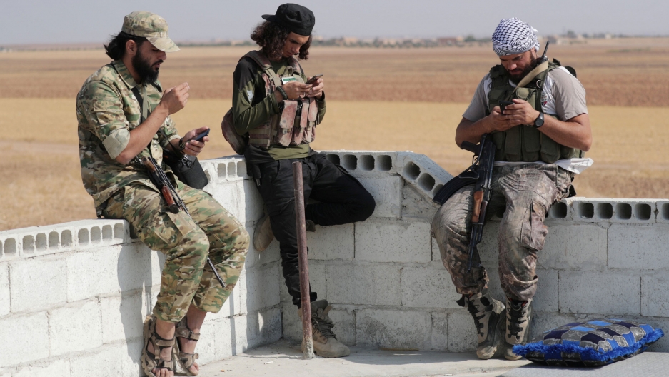 Turkey-backed Syrian rebel fighters use their mobile phone near the border town of Tal Abyad, Syria, Oct. 22, 2019. Three men are shown, two wearing beards and all of them are looking down at their mobile phones.