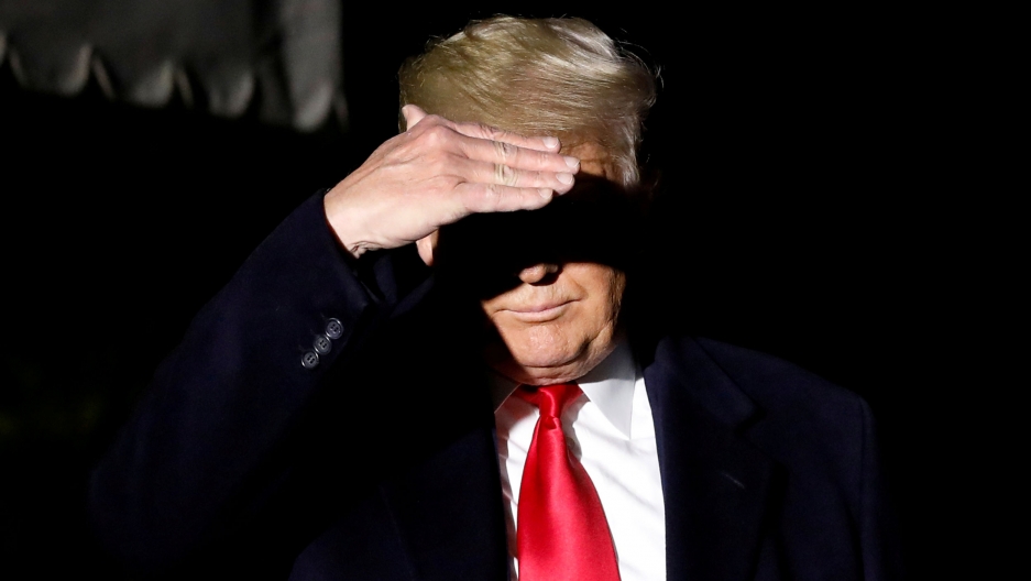 President Donald Trump covers his face from TV lights as he walks toward the media on the South Lawn of the White House, Nov. 2, 2019. President Donald Trump is shown wearing a suit and red tie and using his hand to cover his eyes.