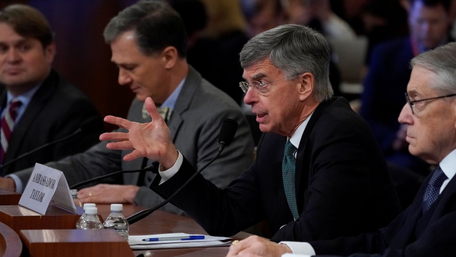 Ambassador William Taylor, the top US diplomat in Ukraine, testifies during the House impeachment inquiry hearing into US President Donald Trump, Nov. 13, 2019. Ambassador William Taylor is shown from the side, sitting at a table with his right hand outstretched.