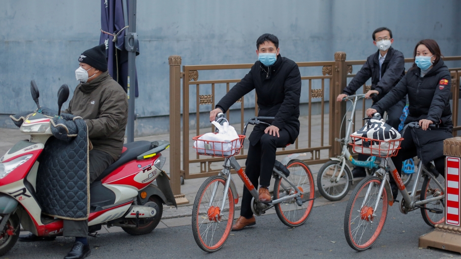 People on bicycles and electric scooters wear masks as they wait at a traffic light after a yellow alert was issued for smog in Beijing, China, on Nov. 14, 2018. Three people are shown riding bicycles along with one riding a scooter and all wearing face masks.