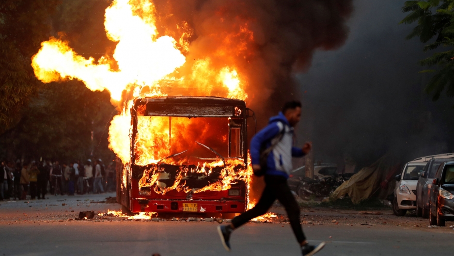 A man runs past a burning bus that was set on fire by demonstrators during a protest against a new citizenship law, in New Delhi, India, Dec. 15, 2019. A man is shown in a blur running past the front of a bus on fire with large explosive-looking flames.