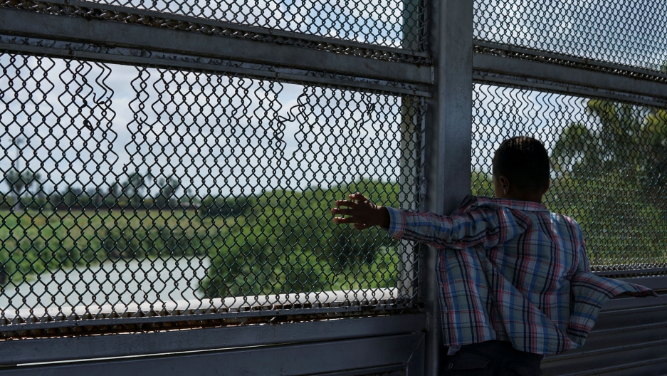 A young boy is shown with a plaid shirt and looking through a fence. A young asylum seeker looks at the Rio Grande in Matamoros, Mexico.